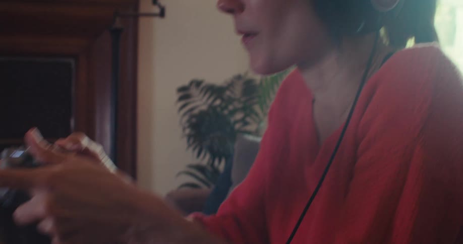 A woman in a red sweater wearing headphones sits on a sunlit couch at home excitedly playing video games