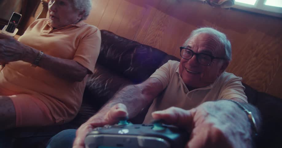 Two elderly people joyfully play video games on a couch in a woodpaneled living room