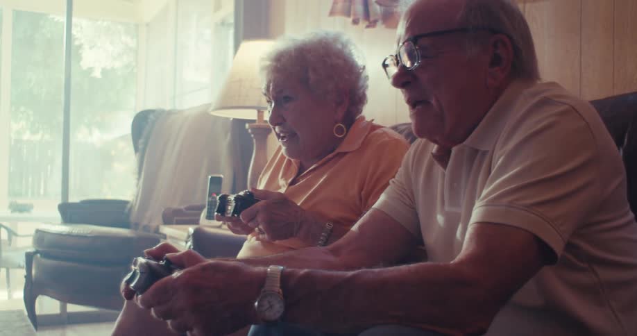 An elderly man and woman sit on a gray couch both holding game controllers with focused expressions as they play
