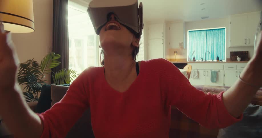 Lady moves her head and hands around while seated on the couch indoor at daytime with virtual reality headset on