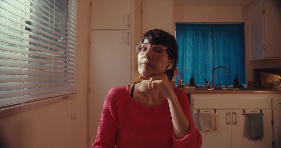 A woman in a red sweater sitting in her kitchen with an earbud in puts on her glasses while looking off screen