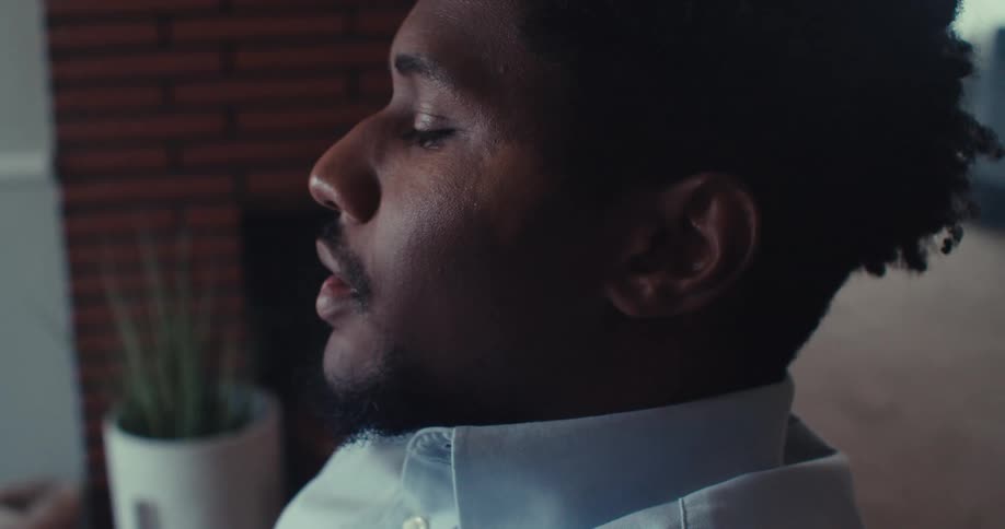 A young black man operates a laptop then sits back looking tired in his office