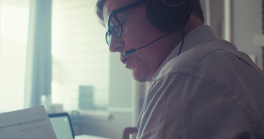 A young man wearing a headset looks at stock prices holds a stock report and talks on a video call on his laptop