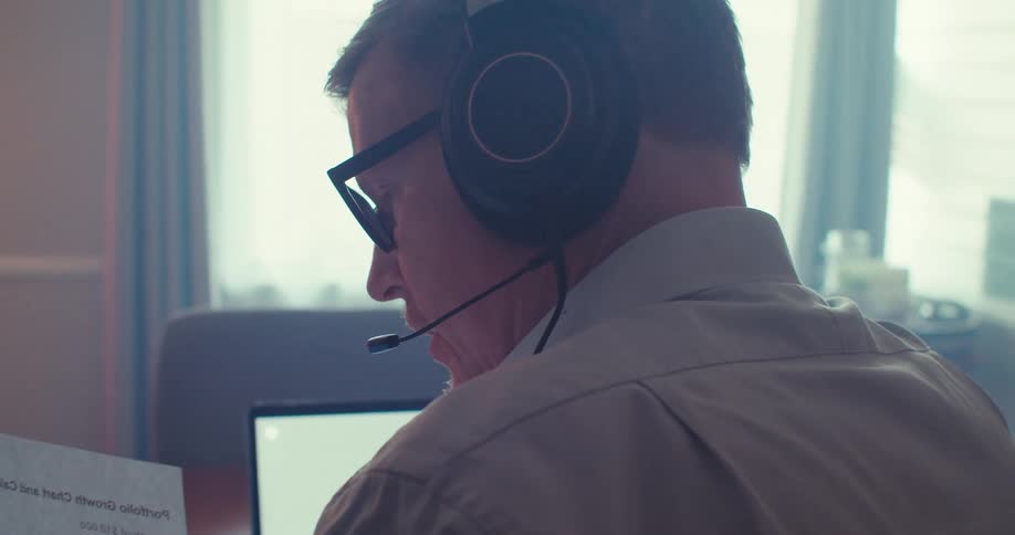 A man sits in an office looking at a document in front of his laptop while speaking into the microphone on his headset around midday