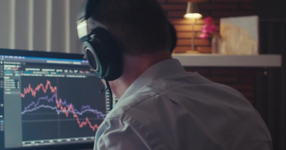 A man wearing headphones yells into a call while analyzing a stock market chart on his screen