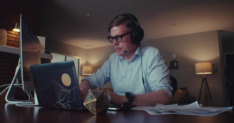 A man in headphones intensely works on his laptop while analyzing documents at a dimly lit desk