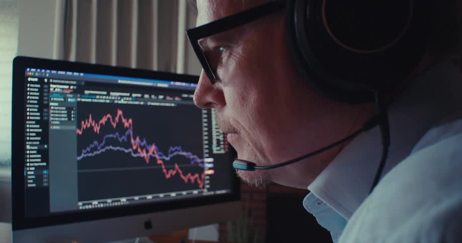 A middleaged man wearing a headset looks at stock prices on a monitor while speaking into the headset microphone