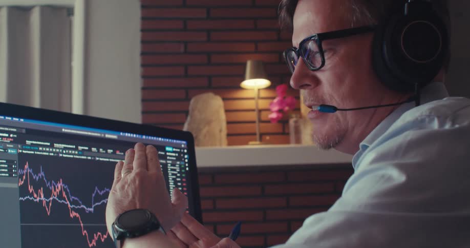 A man wearing a headset with a mic uses hand gestures while explaining during a video call a display monitor in front of him shows stock prices and a laptop sits on the left side of the desk