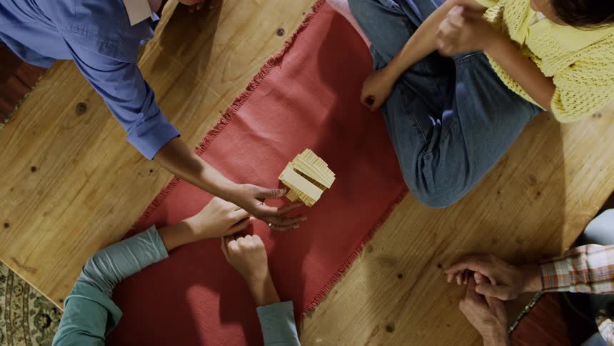 An overhead view captures four peoples hands around a wooden block game on a red mat placed on a wooden floor