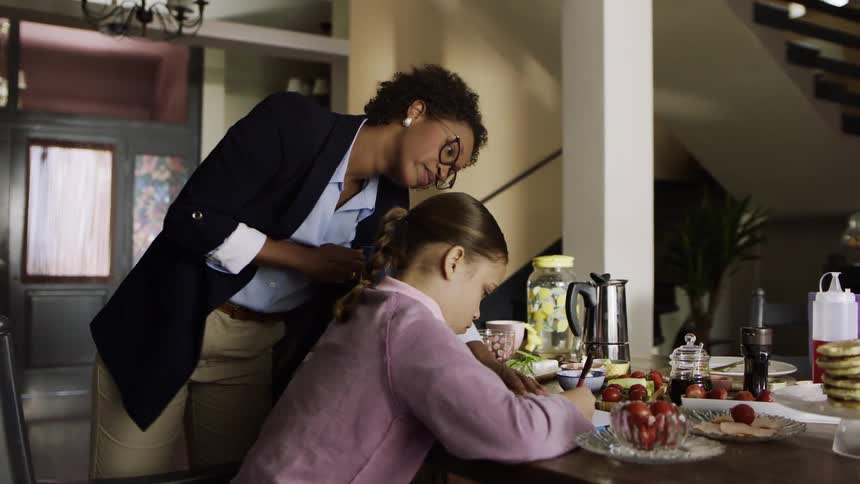 A mother watches her daughter do homework at the table full of food