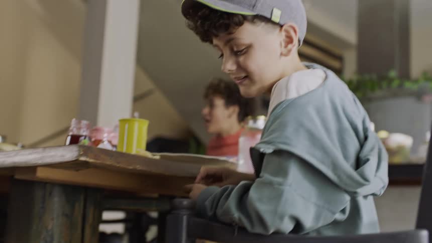 Boy laughs after a dog jumps to eat the food in his hand while seated at the dining table