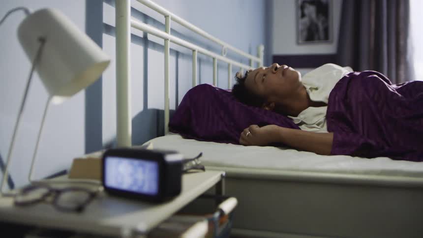 A woman lies in bed reaches to turn off a 630 alarm clock then stands up from the bed