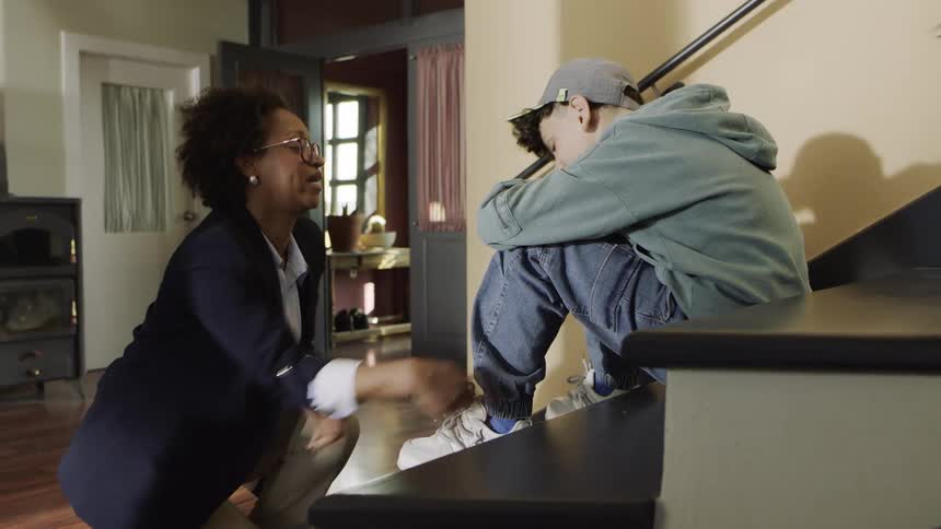 A woman ties a boys shoe as he sits quietly on the stairs