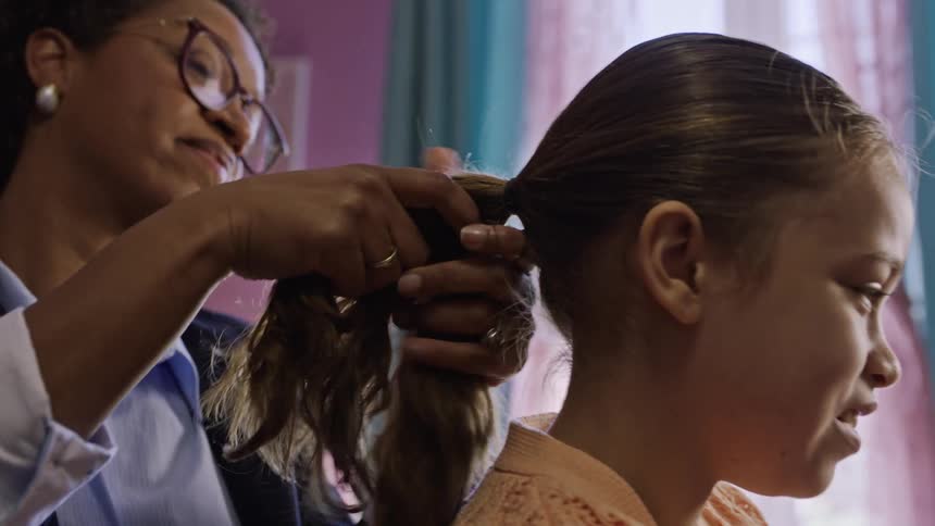 Mother ties a young girls hair into a ponytail in a warmly lit room