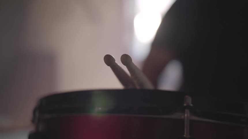 Drumsticks rest on top of a red black rimmed snare drum in a room with natural sunlight coming through a window in the background