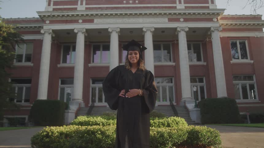 A proud female graduate in a cap and gown smiles while standing in front of a stately brick administration building on a college campus