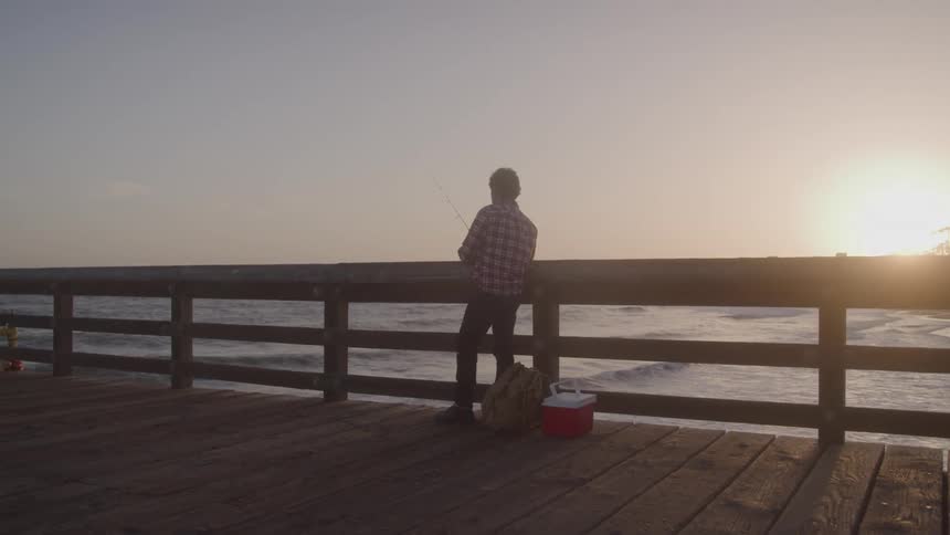 A lone man is silhouetted against the bright sun as he fishes from a wooden pier with his cooler and backpack resting beside him