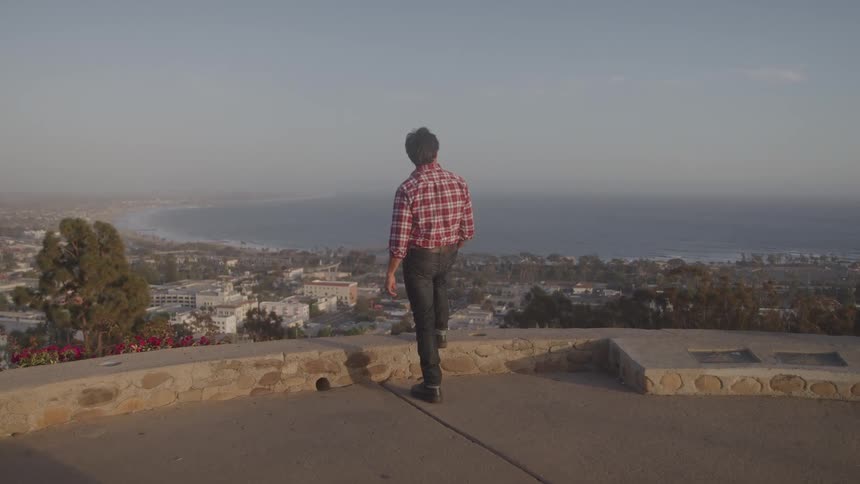 Man in a red plaid shirt stands overlooking a coastal city from a scenic viewpoint