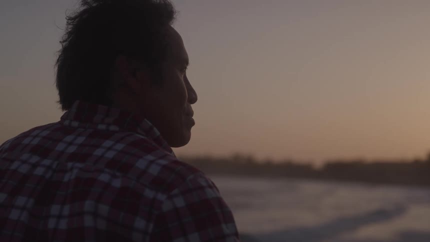 A man in a plaid shirt stands by the ocean at sunset looking toward the waves with a coastline in the distance