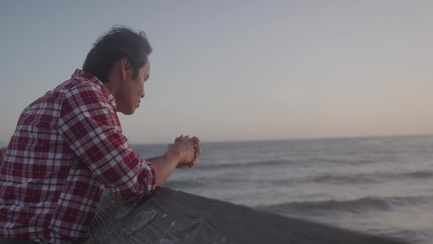 A man in a plaid shirt leans on a wooden railing his hands clasped together as he looks out at the ocean