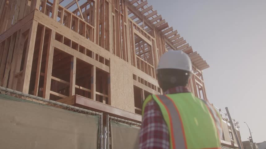 A construction worker wearing a hard hat and highvisibility vest observes a wooden building framework under construction
