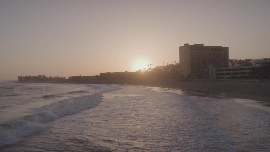 From a pier waves crash on a beach with buildings in the background as the sun sets and people are visible on the sand