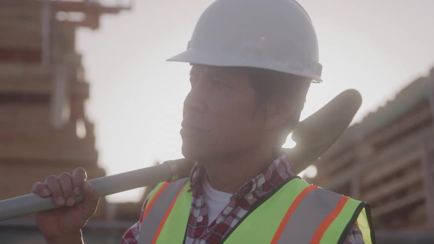 A construction worker stands at a construction site holding a shovel wearing a helmet and a safety green vest