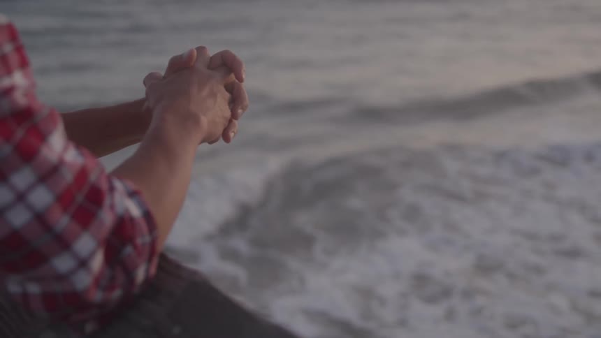 A person in a red and white checkered shirt stands near the shoreline clasping their own hands in a pensive meditative posture as waves roll in front