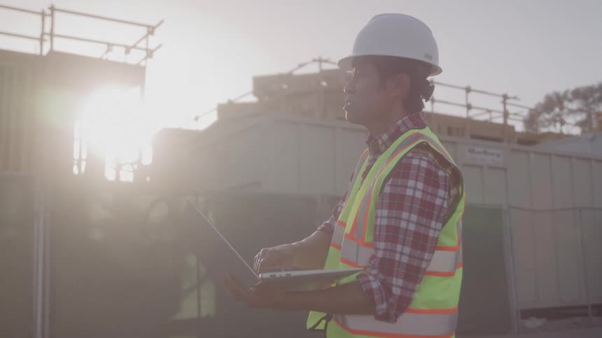 A construction worker is inspecting a site while working on a laptop