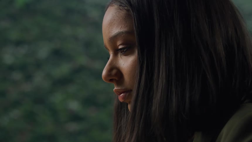 A person with long dark hair takes a bite from a red apple in front of dense green foliage