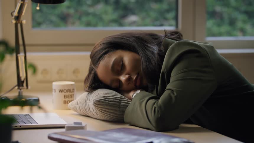 A young woman laying her head on a pillow that is on an office desk and she starts to sleep