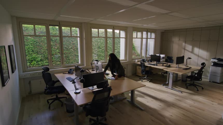A woman walks into an empty office around midday sits on a chair and starts working on a computer