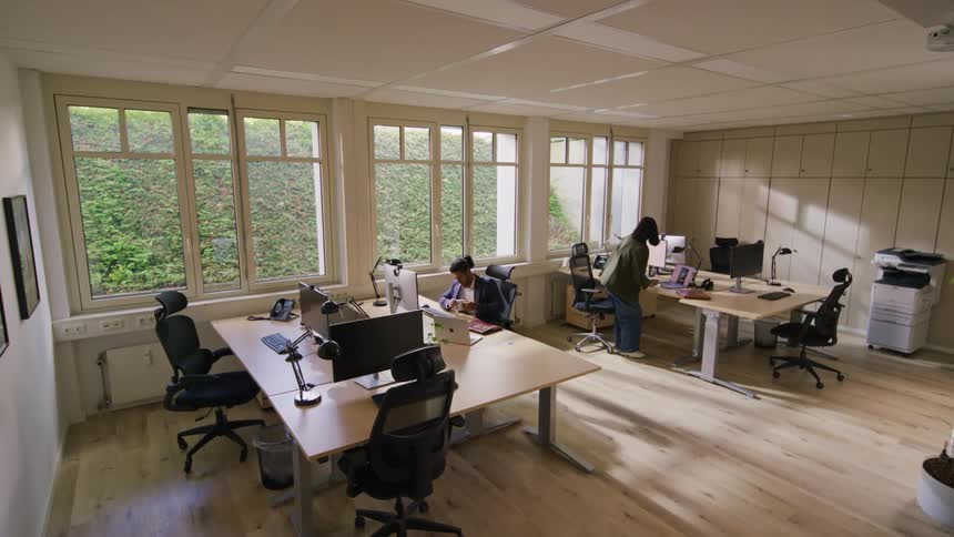 Two people work on laptops at a wooden desk in a modern office with large windows and several vacant workstations