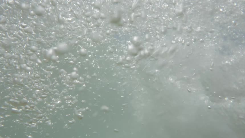 Shot from slightly under the oceans surface the scene fills with a turbulent rush of bubbles as a wave flows by overhead