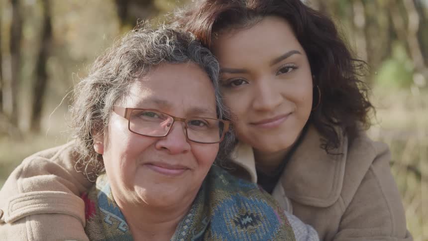 Elderly woman and her granddaughter smile while the younger woman hugs the older one outdoors near the woods