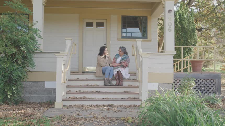 An elderly woman sitting on the steps of a porch of a house next to a younger woman and they are talking