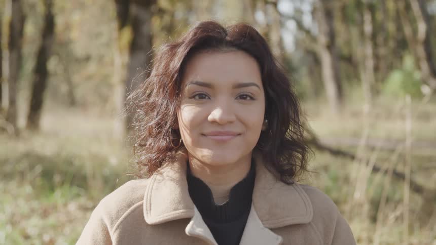 A woman stands outdoors in a forest clearing smiling calmly at the camera