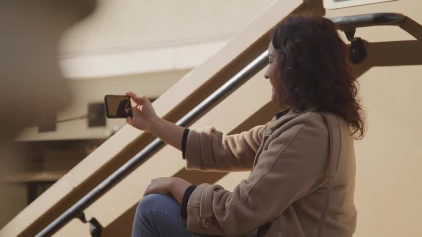 Woman sitting on stairs waving while video calling someone on her phone