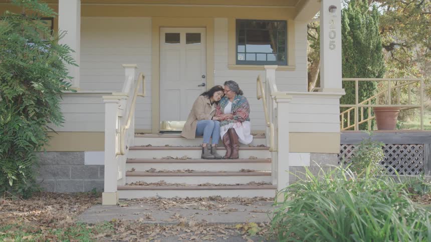 Woman lovingly hugging an elderly woman as they sit outside a house