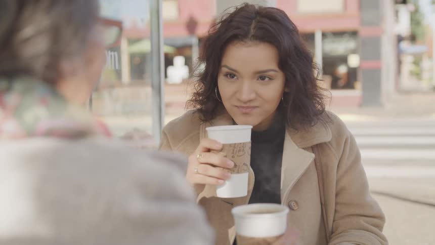 Two women sit at an outdoor caf holding coffee cups and engaged in conversation