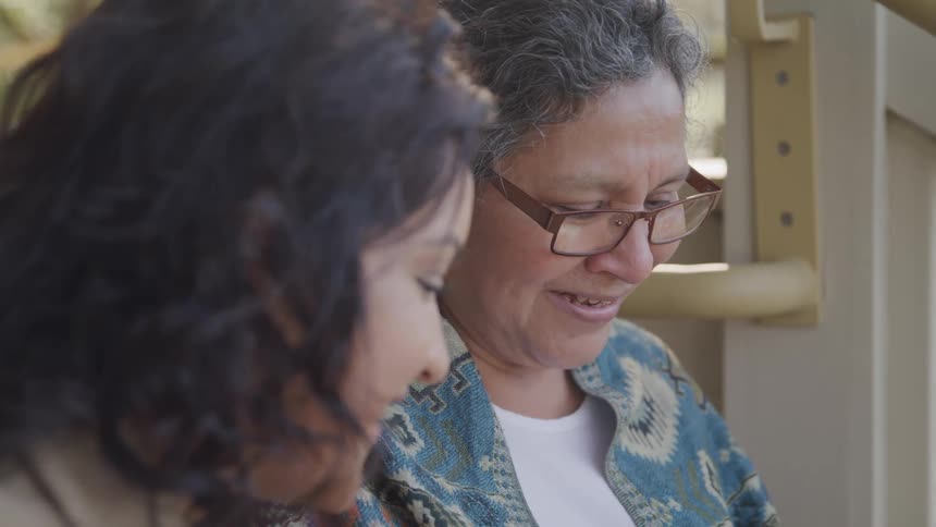 A woman with graying hair and glasses is shown in a close view smiling slightly while talking to another woman next to her