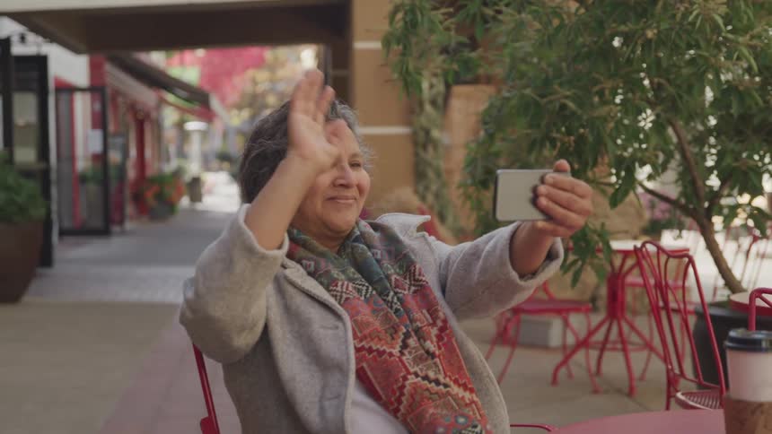 An elderly woman is smiling and waving while video chatting on her phone at an outdoor caf