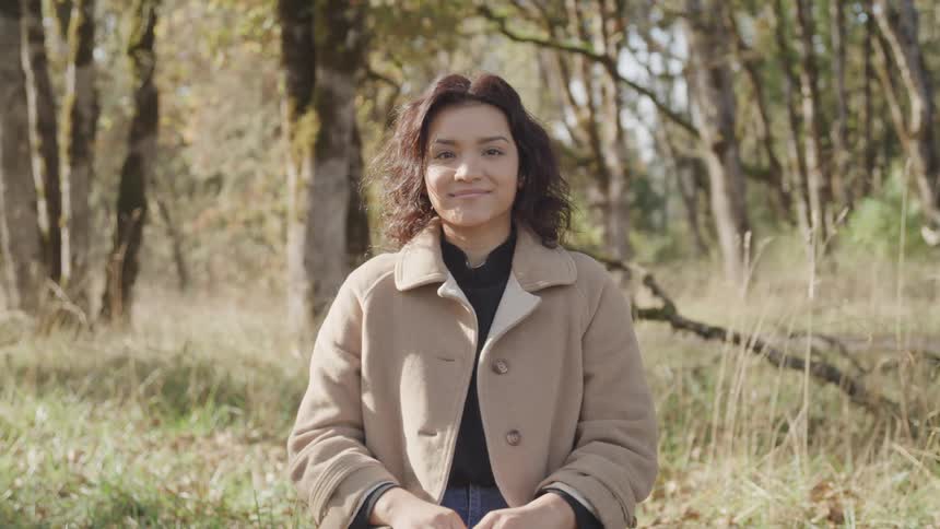 A portrait of a woman who is smiling and looking forward and she is wearing a brown coat and there is a forest behind her