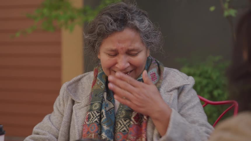 An elderly woman is laughing heartily while sipping coffee at an outdoor caf