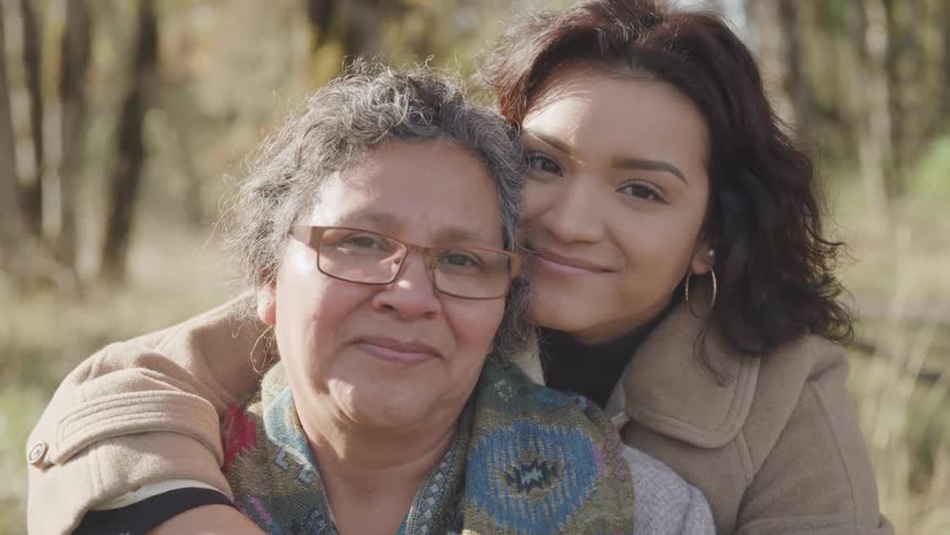 Two women are wearing warm clothes and are hugging each other while looking forward happily