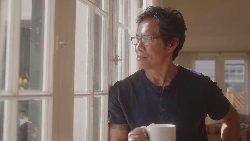 A man stands by a window holding a coffee mug gazing outside thoughtfully