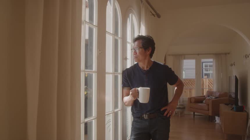 A man stands by a large window holding a coffee mug gazing outside in a sunlit living room