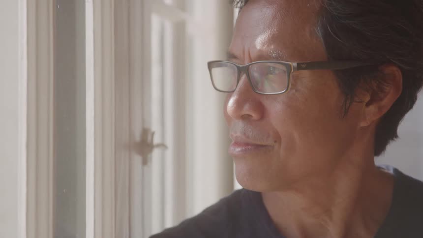 A man with glasses looks thoughtfully out a window in soft natural light