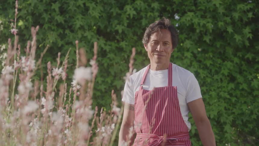 Man in red apron stands in sunny garden with wildflowers