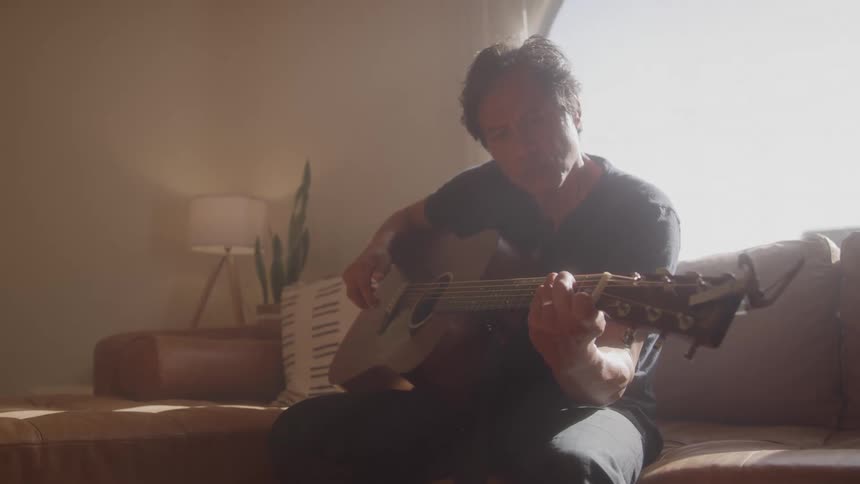 A man plays acoustic guitar while sitting on a couch in soft natural light
